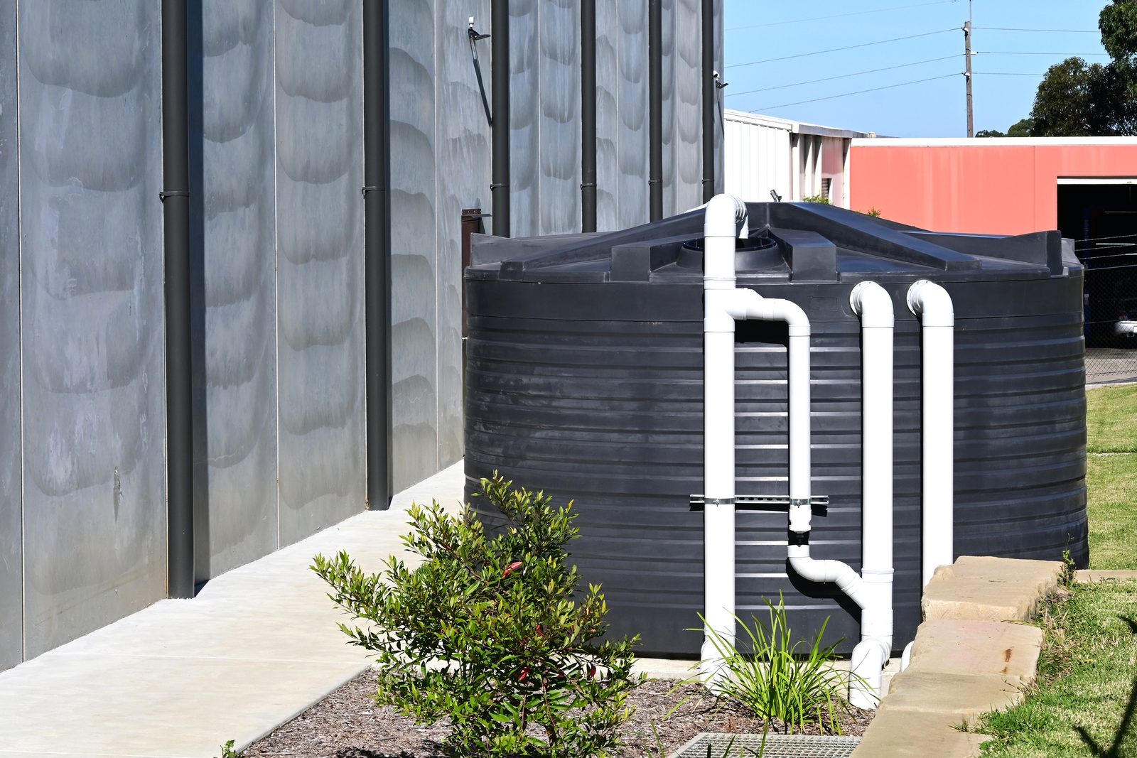 Black poly rainwater tank with white PVC inlet pipes beside a Colorbond wall